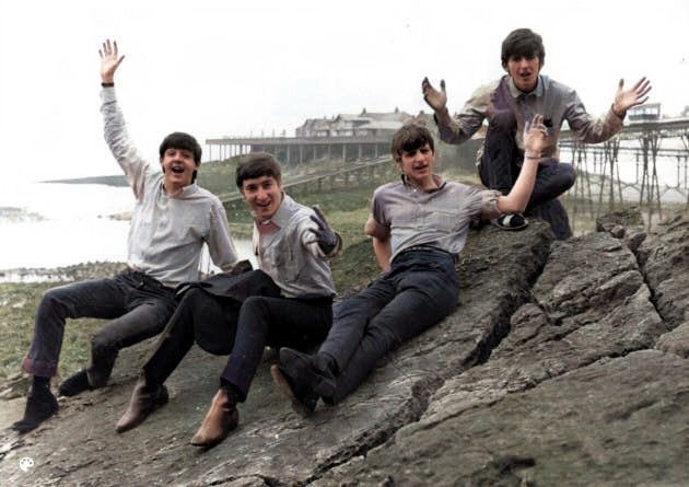 Colour picture of al four of The Beatles sitting on a rock in Weston-super-Mare with Birnbeck Pier in the background